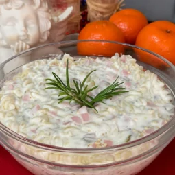 Staklena zdjela puna brze salate s tjesteninom, šunkom, kukuruzom i kremastim umakom, ukrašena grančicom ružmarina, u pozadini nekoliko mandarina