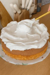 Zlatna špatula širi sloj bijelog pjenastog fila preko čokoladne torte na drvenoj radnoj ploči s kuhinjskim priborom u pozadini.