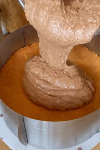 Gusta čokoladna krema izlijeva se iz posude na zlatno pečenu biskvitnu podlogu unutar metalnog kalupa za tortu.