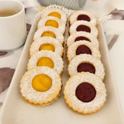 Linzer keksi posuti šećerom u prahu, punjeni pekmezom od marelice i maline, posluženi na tanjuru uz šalicu kave