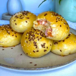 Mekani pizza sendviči iz pećnice punjeni šunkom, sirom i kečapom, posuti sezamom i lanom, servirani na tanjuru u minimalističkoj kuhinji.