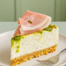 Komad slanog cheesecakea s ricottom, pistacijama i pršutom poslužen na tanjuru u minimalističkoj zelenoj kuhinji.