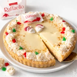 Raffaello tart bez pečenja s kokosom i bijelom čokoladom, kremasti kolač s izvađenim komadom na bijelom tanjuru
