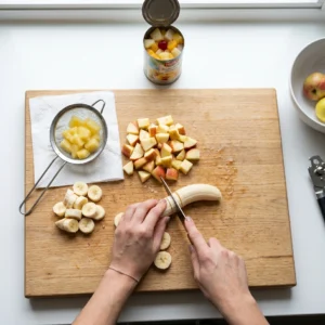 Rezanje banane na drvenoj dasci uz ocijeđeni ananas, kockice jabuke i konzervu voćnog koktela